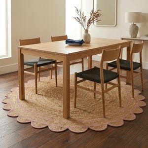 May include: A light wood dining table with four matching chairs. The chairs have black woven seats. The table sits on a round jute rug with a pink scalloped edge. A vase with dried flowers sits on the table.