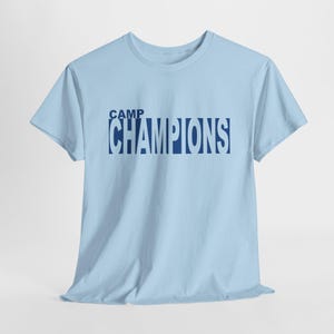 May include: A light blue t-shirt with the text "CAMP CHAMPIONS" printed in navy blue.