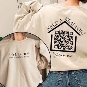 Peut inclure: Sweat-shirt couleur sable avec le texte "NEED A REALTOR?" et un code QR. Le sweat-shirt comporte également le texte "Scan me" et "SOLD BY @custom text here".