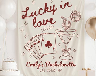 May include: A cream-colored fabric banner featuring dark red line art. The text reads Lucky in love EST.2025, Emily's Bachelorette, and LAS VEGAS, NV. Graphics include a royal flush of playing cards, a martini glass with boots, and a disco ball.