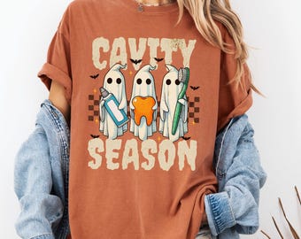 May include: A rust-colored t-shirt featuring a graphic of three white ghosts. One ghost holds a blue toothpaste tube, one holds an orange tooth, and one holds a green toothbrush. The text reads CAVITY SEASON, surrounded by small black bats.