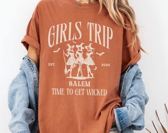 May include: A burnt orange t-shirt with a graphic of three witches and the text "Girls Trip Est. 2024 Salem Time to Get Wicked".