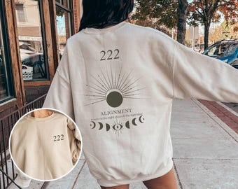 May include: A beige sweatshirt with a graphic design featuring a sun, moon, and eye with the text "Alignment You're in the right place at the right time" and the number "222".