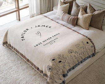 May include: A cream-colored woven throw blanket featuring black line art of wildflowers along the bottom edge. The text reads Taylor + James, a heart symbol, and the phrase Love Never Fails Corinthians 13:8. The blanket has a fringed edge.