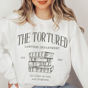 May include: White sweatshirt with the text "THE TORTURED LAWYERS DEPARTMENT EST 2025" and a drawing of a stack of books. The text below the drawing says "All's fair in love and litigation."