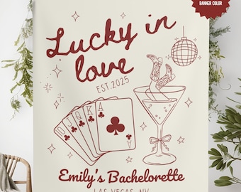 May include: A white banner with red text and illustrations. The text reads "Lucky in love EST. 2025" and "Emily's Bachelorette LAS VEGAS, NV". The illustrations include a martini glass with a pair of legs in cowboy boots sticking out, a disco ball, and a hand of playing cards.