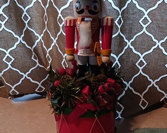 May include: A wooden nutcracker doll in a red and gold uniform stands on a red drum with gold trim and a green base. The drum is surrounded by a green and red Christmas wreath with pine needles, berries, and pinecones.
