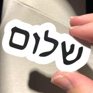 Może przedstawiać: Biała naklejka z hebrajskim słowem "Shalom" w kolorze czarnym, trzymana między palcami. Naklejka ma białą obwódkę i znajduje się na neutralnym tle.