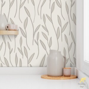 Op de afbeelding: Een witte muur met een grijs bladmotief behang. Een witte kan en een kleine grijze container staan op een houten oppervlak voor de muur.