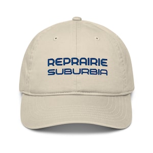 Może przedstawiać: Beżowa czapka z daszkiem z haftowanym napisem "REPRAIIRIE SUBURBIA" w kolorze niebieskim.