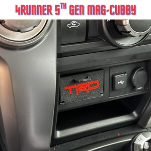 May include: A black magnetic storage cubby insert for a 5th generation 4Runner centre console, featuring a textured surface and a red TRD logo. The text 4RUNNER 5TH GEN MAG-CUBBY is displayed above the console storage compartment.