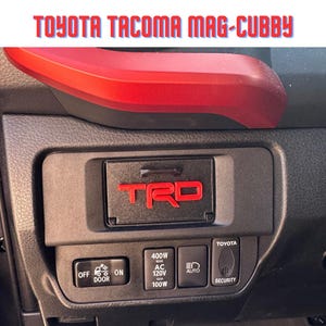May include: Black dashboard component with red accents and the text "TOYOTA TACOMA MAG-CUBBY." Features a "TRD" logo and control buttons labeled "OFF DOOR ON," "400W AC 120V 100W," "AUTO," and "SECURITY."