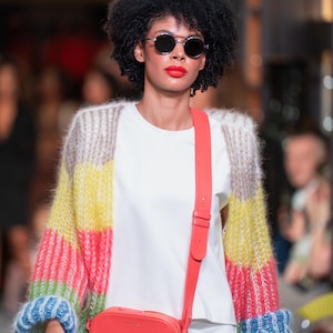 May include: A model on a catwalk wearing a white t-shirt, round sunglasses, and a colourful striped cardigan with blue cuffs. A coral crossbody bag is worn over the shoulder. The model has red lipstick.