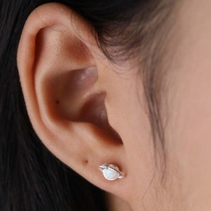 May include: A close-up of a person's ear with a silver Saturn stud earring featuring a white opal stone.