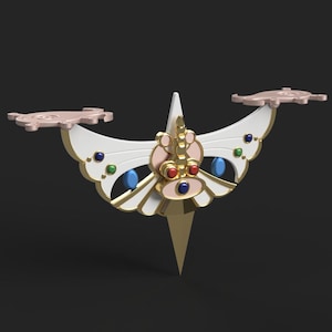 Op de afbeelding: Een gouden en wit vlindervormig decoratief object met rode, blauwe en groene edelstenen. Het object heeft een puntige onderkant en twee vleugels met een roze en wit ontwerp.