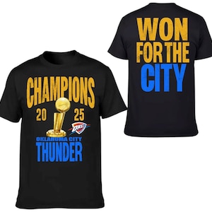 Op de afbeelding: Zwarte t-shirts met gouden en blauwe tekst en afbeeldingen. De voorkant van het shirt zegt "CHAMPIONS 2025" met een trofee-afbeelding en "OKLAHOMA CITY THUNDER". De achterkant van het shirt zegt "WON FOR THE CITY".