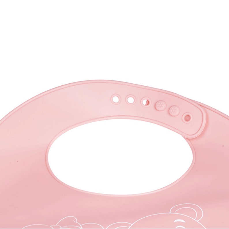 May include: Pink silicone baby bib with a bear design and adjustable neck strap. The bib has a pocket to catch food and a soft, flexible texture.