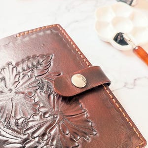 Peut inclure: Un carnet en cuir marron avec un motif floral en relief et une fermeture à bouton argenté.