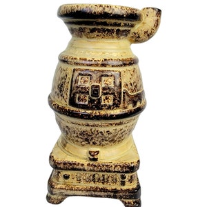 May include: A ceramic stove-shaped vase with a brown and yellow glaze. The vase has a small spout on the side and a decorative design on the front.