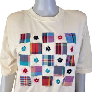 May include: A white t-shirt with a patchwork design featuring a grid of colorful plaid squares with embroidered flowers.