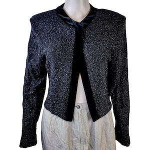 May include: A black and silver textured jacket with a black velvet trim and collar. The long-sleeved jacket has a button closure and is displayed on a mannequin. The jacket is paired with light-colored pants.