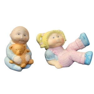 May include: Two porcelain Cabbage Patch Kids dolls. The doll on the left is wearing a blue outfit and holding a brown teddy bear. The doll on the right is wearing a pink and blue outfit and has blonde hair.