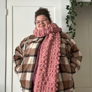 May include: A person wearing a brown and white checked shirt with a pink crocheted scarf. The scarf has a textured, puffy design.