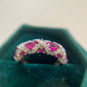 May include: A silver band ring with a row of oval-shaped ruby stones set in a pave setting with smaller diamonds.
