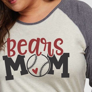 Puede incluir: Una camiseta raglán con temática de béisbol con las palabras "bears MOM" en rojo y negro. La "O" en MOM es una pelota de béisbol con un corazón y detalles de costura. La camiseta tiene un cuerpo crema y mangas grises.