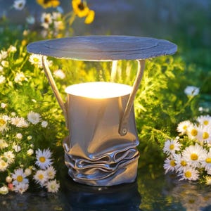 May include: A crushed metal can with a light inside, used as an outdoor lamp. The lamp is on a slate base and surrounded by white flowers.