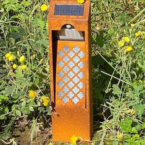 Peut inclure: Un lampadaire de jardin rectangulaire à énergie solaire, d'aspect vieilli. Il est doté d'un panneau solaire sombre sur le dessus, d'une section de petites lumières LED et d'un panneau décoratif avec des découpes en forme de quadrilobe argenté. Entouré d'herbe verte et de fleurs sauvages jaunes.