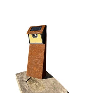 Può includere: Un'apparecchio di illuminazione per esterni a energia solare con un palo metallico rettangolare color ruggine. La luce ha un pannello solare nero e un alloggiamento della luce giallo e nero. L'apparecchio è montato su una base di legno stagionato.