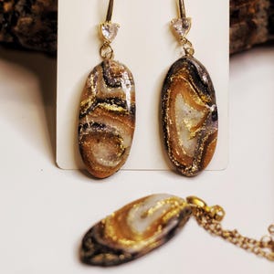 Könnte beinhalten: Ein Schmuckset mit Ohrringen und einer Anhängerkette. Die Ohrringe haben goldfarbene Haken mit herzförmigen, klaren Steinen. Die ovalen Ohrringe und der Anhänger haben ein marmoriertes Design in Braun-, Gold-, Schwarz- und Weißtönen.
