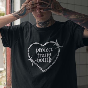 以下が含まれることがあります： ハートが有刺鉄線で囲まれた白いグラフィックが描かれた黒いTシャツ。ハートの中のテキストは「protect trans youth」です。