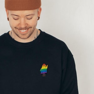 A cute small rainbow ghost waving on the top left pocket of a black sweatshirt