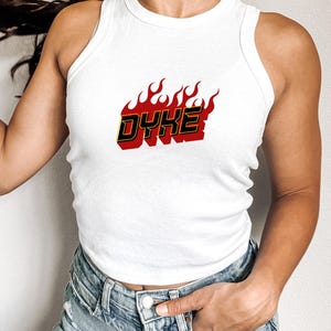May include: White tank top with the word "DYKE" in red and black, surrounded by flames. The text is in a bold, stylized font. The top is sleeveless and made of a soft material.