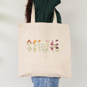 Puede incluir: Una bolsa de tela beige con un diseño de flores coloridas en forma de arcoíris. Las flores están dispuestas en una línea a través de la bolsa.