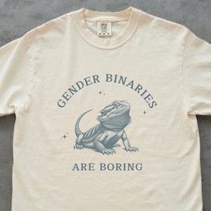 Op de afbeelding: Een beige T-shirt met een blauwe afbeelding van een baardagame en de tekst "Gender Binaries Are Boring".