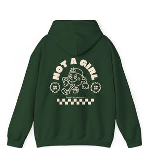 May include: A dark green hoodie with a white graphic print on the back. The graphic features a cartoon character walking with the text "NOT A GIRL" in a circle around the character. The text "Gender is a construct" is printed below the graphic.