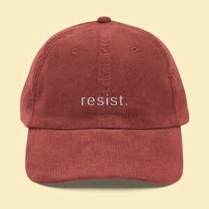 May include: A maroon corduroy baseball cap with the word "resist" embroidered in white. The cap has a curved peak and a rounded top.
