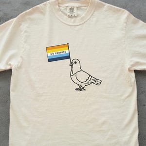 Op de afbeelding: Crèmekleurig T-shirt met een zwarte omtrek tekening van een duif die een vlag vasthoudt. De vlag heeft horizontale strepen in oranje, geel, blauw en wit, met de tekst "NO CRUSHES". Het shirt heeft een ronde hals en korte mouwen.