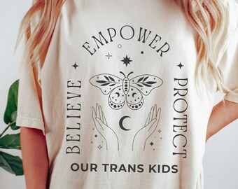 Può includere: Una maglietta color crema con un design grafico nero che presenta una farfalla, mani e una luna crescente. Il testo "EMPOWER BELIEVE PROTECT OUR TRANS KIDS" è stampato sulla maglietta.