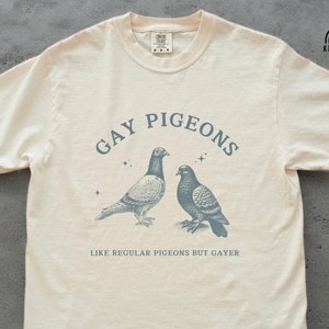 Peut inclure: Un t-shirt crème avec un motif de deux pigeons et le texte "Gay Pigeons Like Regular Pigeons But Gayer".