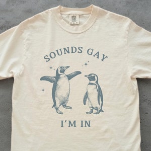 sounds gay i'm in with 2 vintage illustrations of penguins in blue on an ivory comfort colors tshirt