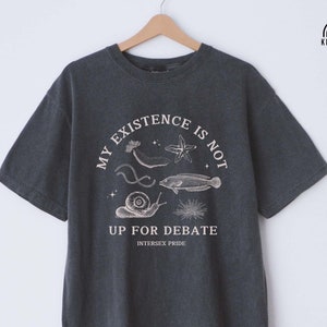 May include: A dark gray t-shirt with a graphic print of a snail, starfish, and fish. The text on the shirt reads "My existence is not up for debate. Intersex Pride."