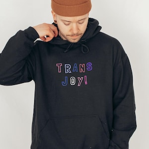 May include: Black hoodie with the text "TRANS JOY!" in a colorful, outlined font. The letters are in shades of pink, blue, and purple. The hoodie has a drawstring and a front pocket.