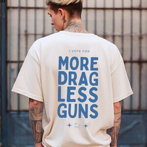 the words 'I vote for more drag less guns' in a stylish typography design on the back of an ivory comfort colors shirt
