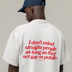 May include: White t-shirt with red text on the back that reads "I don't mind straight people as long as they act gay in public." A person wearing a navy blue baseball cap is in the image.