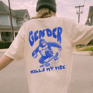 以下が含まれることがあります： 薄いベージュのTシャツに、スケートボードに乗ったゴリラの青いグラフィックがプリントされています。ゴリラの真下に「GENDER KILLS MY VIBE」という青い文字がプリントされています。