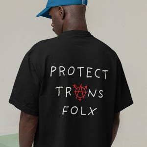 May include: Black t-shirt with the words "PROTECT TRANS FOLX" in white, with a red symbol in the center. A blue baseball cap is worn by the person wearing the shirt.